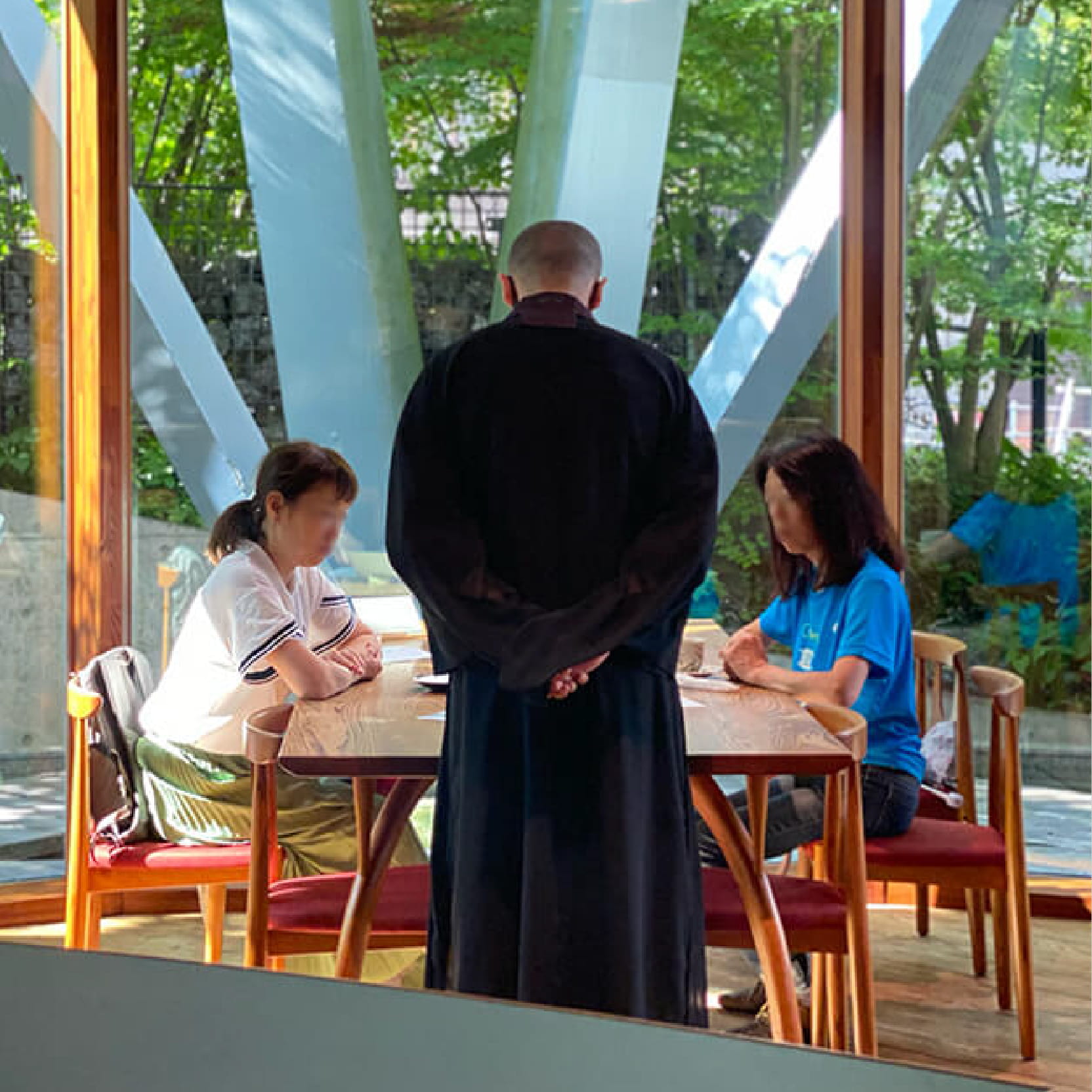 木製テーブルと椅子に座っている二人の女性と、彼女たちの前に立っている僧侶