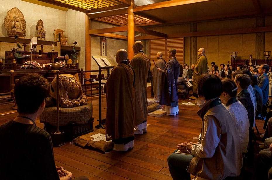 木製の内装が温かみのある仏教寺院の祈りの空間で、僧侶たちが信者の前で儀式を執り行っている様子、前方には仏像と供物が飾られた祭壇が見える