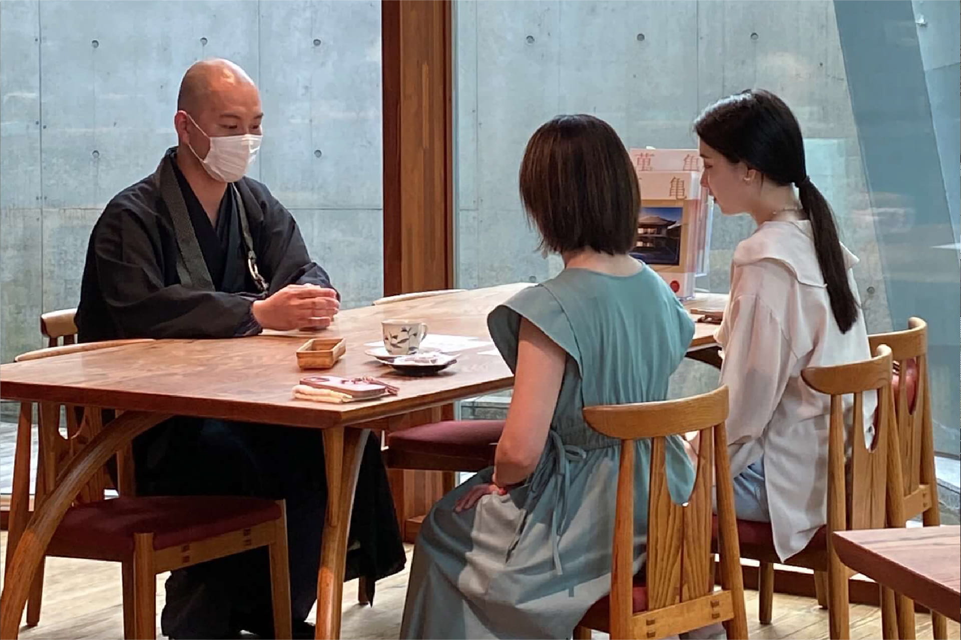 マスクを着用した僧侶が二人の女性と木製テーブルで対面して話している様子